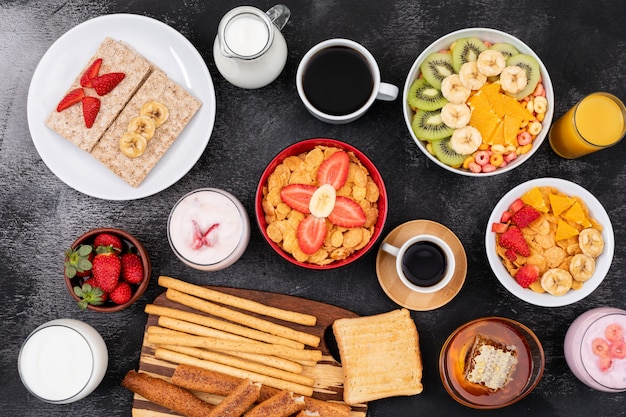 Vista superior del desayuno con frutas, tostadas, copos de maíz, yogurt en superficie negra horizontal