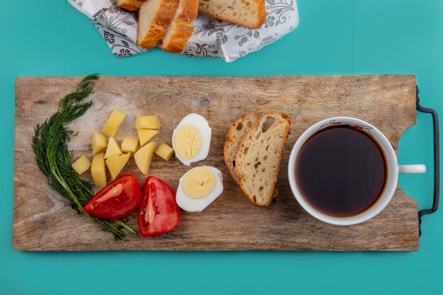 Vista superior del desayuno con huevo, tomate, papa y eneldo con una taza de té en la tabla de cortar y panes sobre fondo azul.