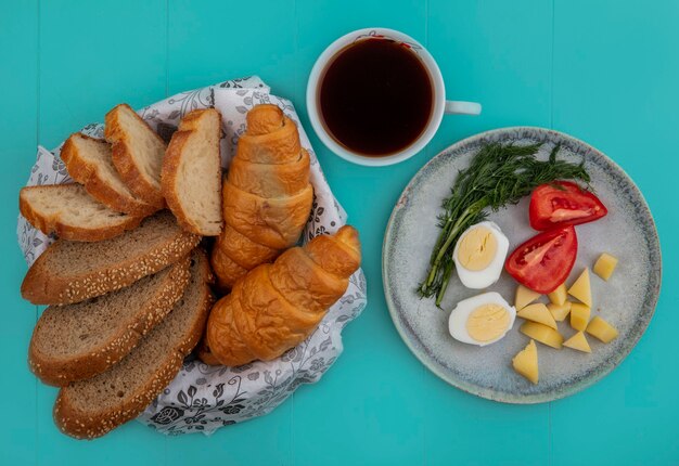 Vista superior del desayuno con huevo, tomate, patata y eneldo con taza de té y panes sobre fondo azul.