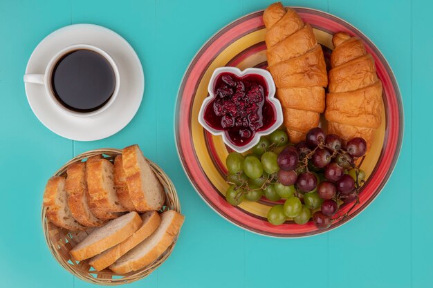 Vista superior del desayuno con mermelada de frambuesa de uva croissant y rebanadas de pan con una taza de té sobre fondo azul.