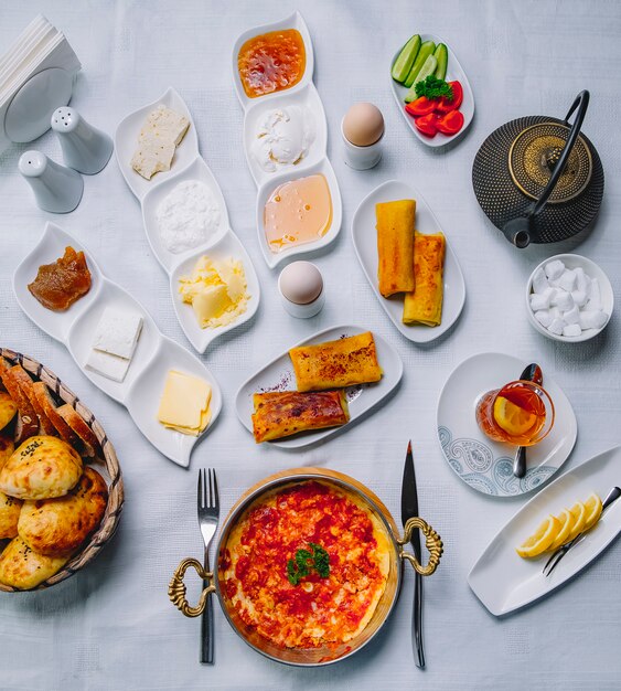 Vista superior desayuno servido mesa huevos revueltos con tomates en una sartén con panqueques queso crema agria mermelada miel y un vaso de té