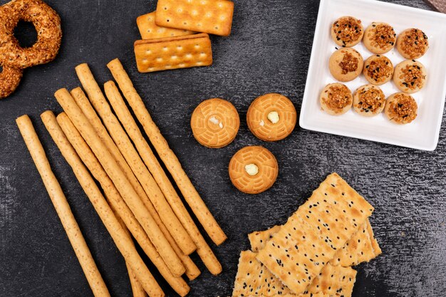 Vista superior de diferentes galletas y palitos de pan en la superficie oscura