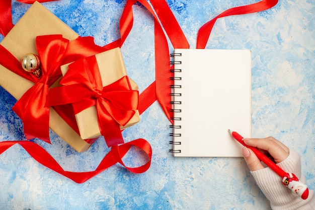 Vista superior de diferentes tamaños de regalos atados con cinta roja, lazo rojo en el bloc de notas en la mano femenina en el cuadro blanco azul