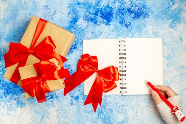 Vista superior de diferentes tamaños de regalos de navidad atados con lazo rojo lazo rojo en el bloc de notas en la mano femenina en la mesa azul