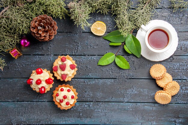 Vista superior de diferentes tartas cono abeto deja juguetes de navidad rodaja de limón una taza de té y galletas en la mesa de madera oscura con espacio de copia