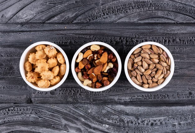 Vista superior de diferentes tipos de aperitivos como nueces y galletas saladas en cuencos en horizontal oscuro