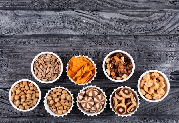 Vista superior de diferentes tipos de bocadillos como nueces, galletas y galletas en tazones con espacio de copia en superficie oscura horizontal