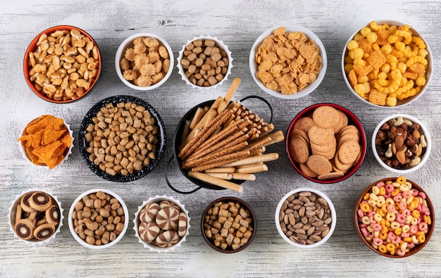 Vista superior de diferentes tipos de bocadillos como nueces, galletas y galletas en tazones sobre superficie de madera blanca horizontal