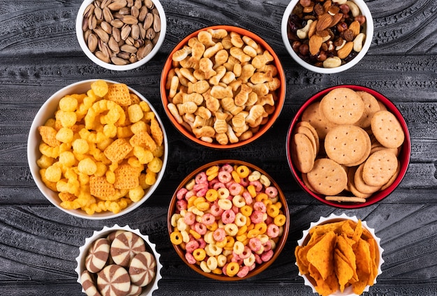 Vista superior de diferentes tipos de bocadillos como nueces, galletas y galletas en tazones sobre una superficie oscura horizontal