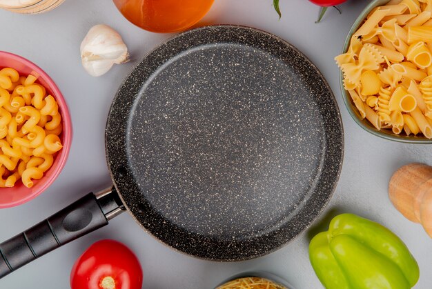 Vista superior de diferentes tipos de macarrones como cavatappi y otros con ajo, tomate, mantequilla, pimienta alrededor de la sartén sobre púrpura