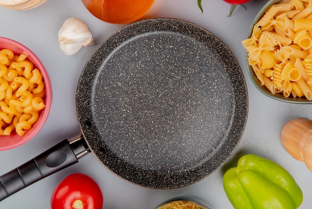 Vista superior de diferentes tipos de macarrones como cavatappi y otros con ajo, tomate, mantequilla, pimienta alrededor de la sartén sobre púrpura