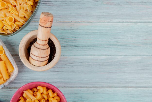 Vista superior de diferentes tipos de pasta como ziti fusilli y otras con semillas de pimienta negra en trituradora de ajo sobre superficie de madera con espacio de copia