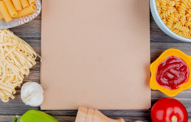 Vista superior de diferentes tipos de pasta como ziti rotini tagliatelle y otros con ajo, tomate, pimiento y salsa de tomate alrededor del bloc de notas en la superficie de madera con espacio de copia