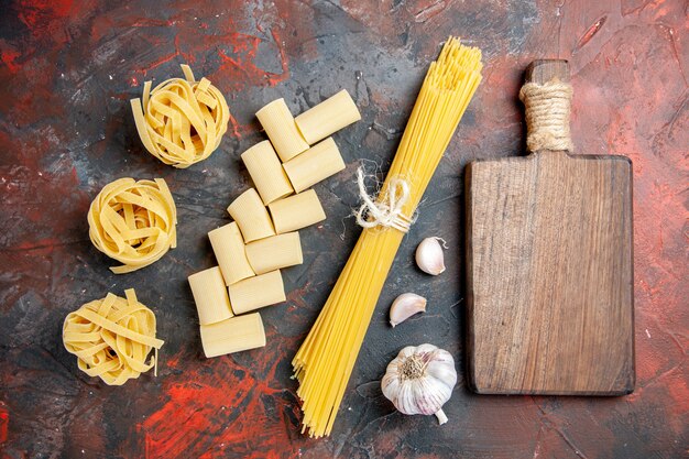 Vista superior de diferentes tipos de pastas crudas y ajo de tabla de cortar de madera sobre fondo negro