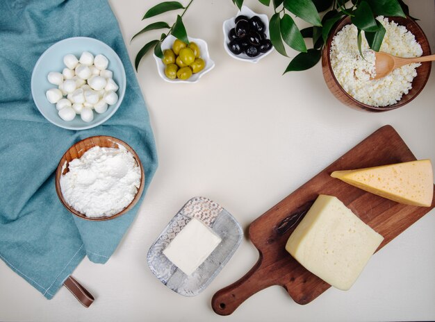 Vista superior de diferentes tipos de queso en tabla de cortar de madera y queso cottage en un tazón de madera con aceitunas en vinagre en mesa blanca