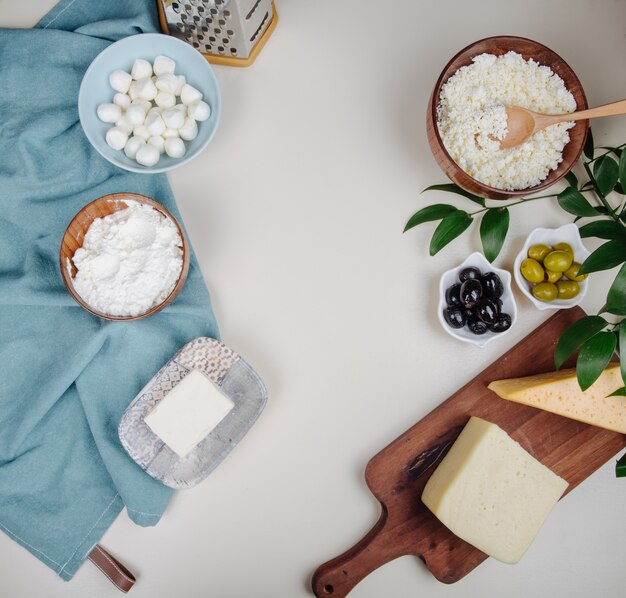 Vista superior de diferentes tipos de queso en tabla de cortar de madera y queso cottage en un tazón de madera con aceitunas en vinagre en mesa blanca