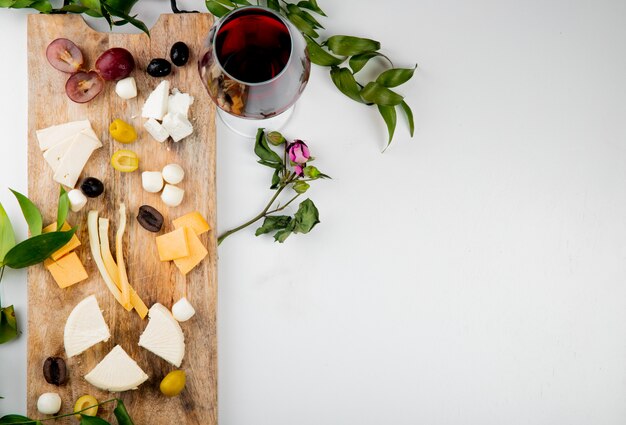 Vista superior de diferentes tipos de queso con trozos de uva aceitunas en tabla de cortar con vino tinto en blanco decorado con flores y hojas con espacio de copia