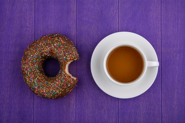 Vista superior de la dona mordida de chocolate con una taza de té sobre una superficie de color púrpura brillante