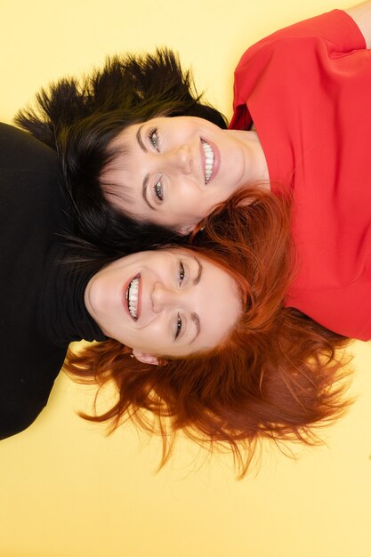 Vista superior de dos mujeres joviales felices en rojo y negro sonriendo felizmente a la cámara en amarillo. Dos mujeres optimistas con cabello rojo y oscuro riendo tirado en el suelo.