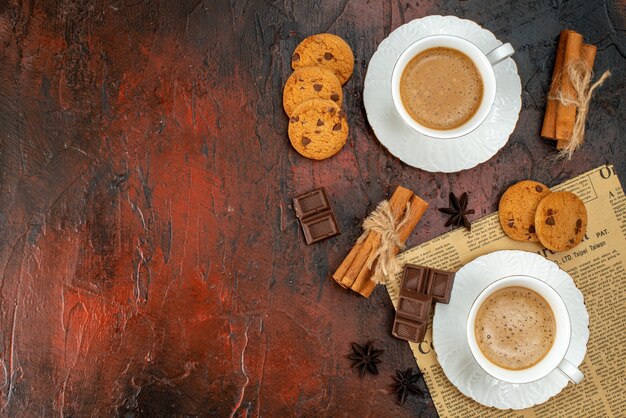 Vista superior de dos tazas de café, galletas, canela, limas, barras de chocolate en un periódico viejo en el lado izquierdo sobre fondo oscuro