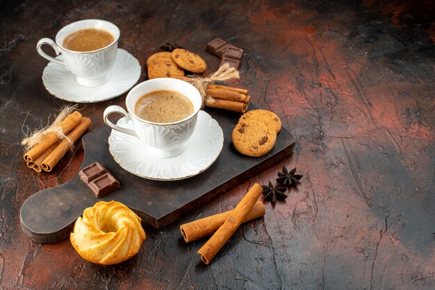 Vista superior de dos tazas de café, galletas, canela, limas, barras de chocolate sobre tabla de cortar de madera sobre fondo oscuro