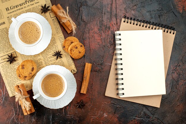 Vista superior de dos tazas de café, galletas, canela, limas en un periódico viejo y cuadernos sobre fondo oscuro