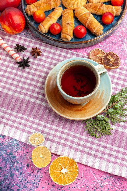 Vista superior dulces deliciosos bagels dentro de la bandeja con ciruelas, melocotones frescos y taza de té en el escritorio rosa claro