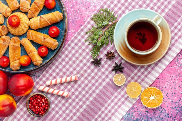 Vista superior dulces deliciosos bagels dentro de la bandeja con ciruelas, melocotones frescos y taza de té en el escritorio rosa claro