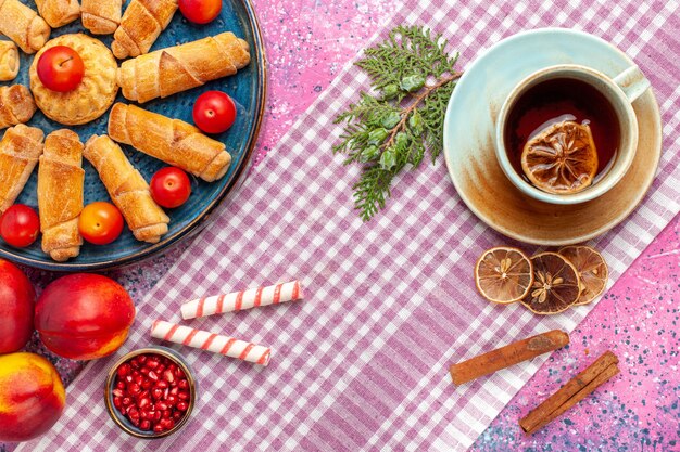 Vista superior dulces deliciosos bagels dentro de la bandeja con ciruelas, melocotones y taza de té en el escritorio rosa claro