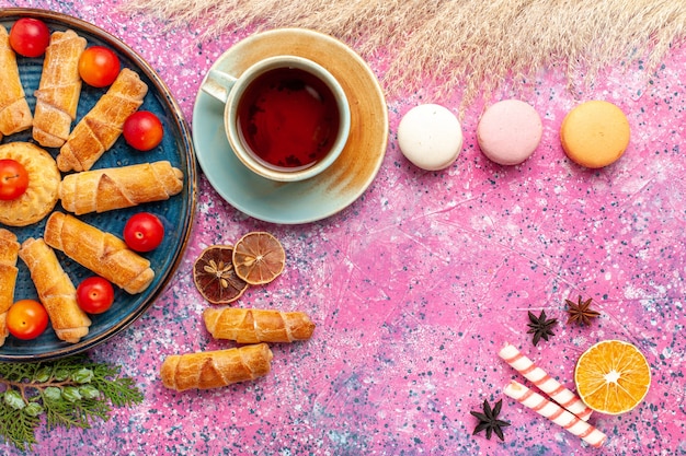 Vista superior dulces deliciosos bagels con macarons franceses de ciruelas ácidas frescas y taza de té en el escritorio rosa claro