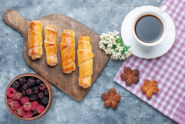 Vista superior dulces deliciosos brazaletes con relleno junto con una taza de café, galletas, bayas en el escritorio de madera gris, azúcar dulce, hornear, pastelería, galleta, galleta