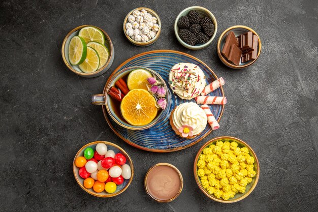 Vista superior de dulces lejanos en la mesa, tazones de fuente de diferentes bayas, chocolate, frutas cítricas, dulces coloridos y apetitosos cupcakes y una taza de té con palitos de limón y canela en la mesa oscura