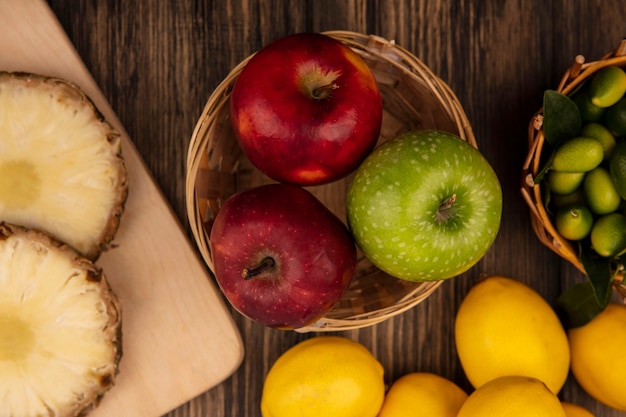 Vista superior de dulces manzanas coloridas en un balde con piñas en una tabla de cocina de madera con kinkans en balde con limones aislado en una superficie de madera