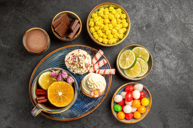 Vista superior de dulces en la mesa cuencos de chocolate frutas cítricas crema de chocolate dulces coloridos y un plato de cupcakes y una taza de té con limón en la mesa oscura