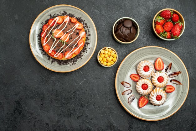 Vista superior de dulces y pastel apetitoso pastel y galletas junto a cuencos de chocolate con nueces de fresa en la mesa oscura