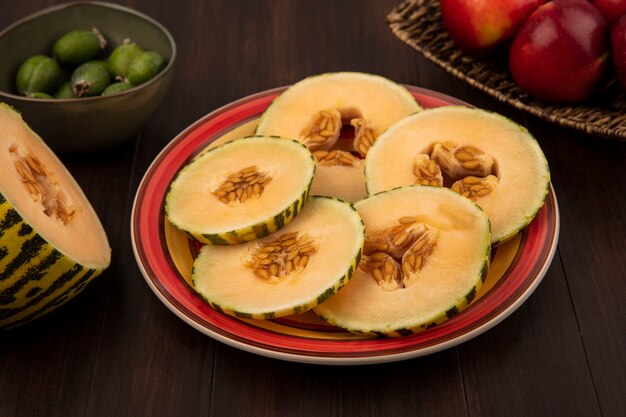 Vista superior de dulces rodajas de melón cantalupo en un plato con feijoas en un recipiente con manzanas en una bandeja de mimbre en una pared de madera