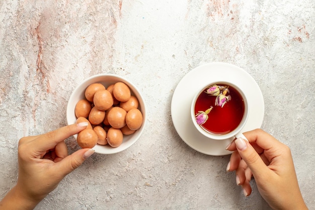 Vista superior de dulces con una taza de té, tazón de dulces y una taza de té en la mano sobre la superficie blanca