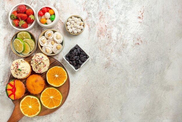 Vista superior de dulces en tazones seis tazones de dulces de bayas y delicias turcas junto al tablero con rodajas de naranja y galletas en la mesa blanca