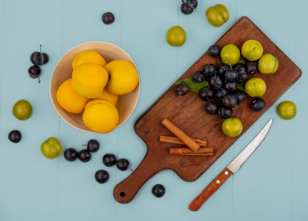 Vista superior de duraznos amarillos frescos en un cuenco con endrinas en una tabla de cocina de madera con ciruelas cereza verde con cuchillo sobre un fondo azul.