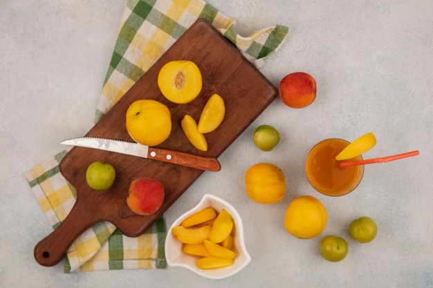 Vista superior de duraznos amarillos frescos en una tabla de cocina de madera con un cuchillo con rodajas de durazno picadas en un recipiente blanco con jugo de durazno en un vaso sobre un fondo blanco.