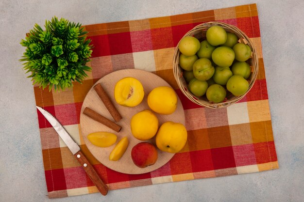 Vista superior de duraznos amarillos sobre una tabla de cocina de madera con ramas de canela con cuchillo con ciruelas cereza verde en un cubo sobre un mantel a cuadros sobre un fondo blanco