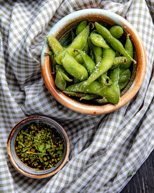 Vista superior de edamame al vapor con sal en un recipiente de arcilla servido con salsa sobre tela escocesa