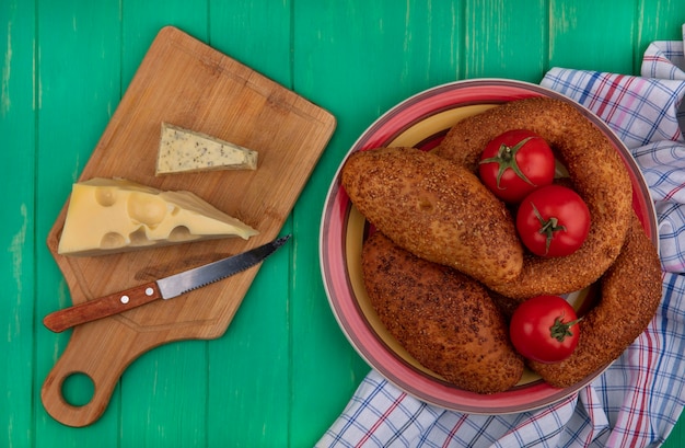 Vista superior de empanadas suaves y de sésamo en un recipiente con tomates frescos sobre un paño con queso sobre una tabla de cocina de madera con cuchillo sobre un fondo de madera verde