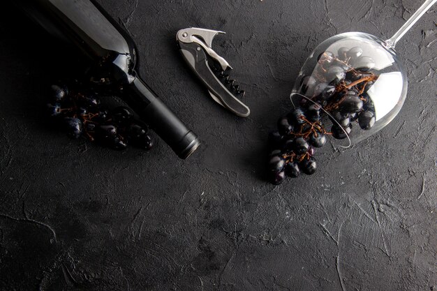 Vista superior encantadoras uvas en copa de vino volcada y abridor de vino de botella de vino en mesa negra