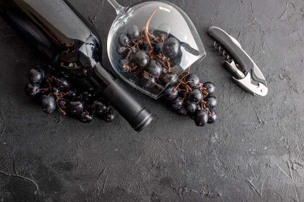 Vista superior encantadoras uvas negras en copa de vino volcada y abridor de vino de botella de vino sobre suelo oscuro