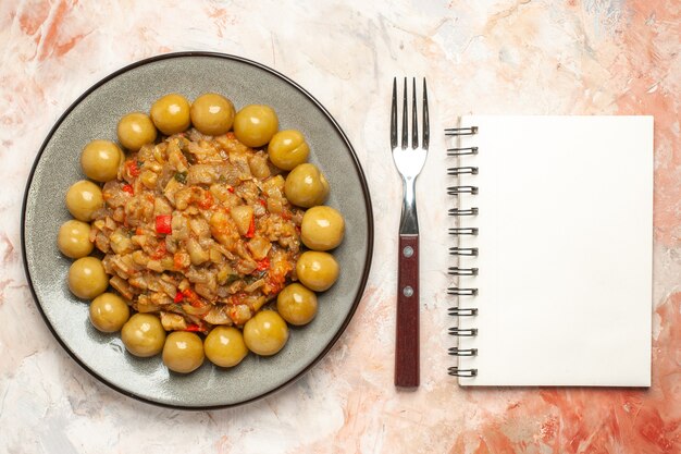 Vista superior de ensalada de berenjena asada y ciruelas en escabeche en un plato tenedor un cuaderno sobre superficie desnuda