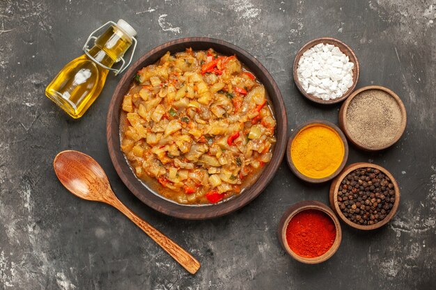 Vista superior de la ensalada de berenjena asada en un tazón, botella de aceite, cuchara de madera y diferentes especias en tazones sobre una superficie oscura