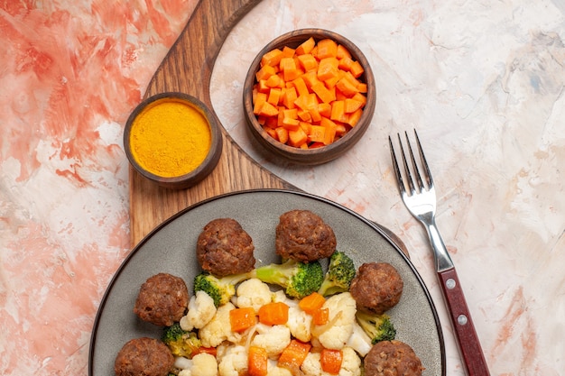 Vista superior de ensalada de brócoli y coliflor y albóndigas en un plato y cúrcuma en la tabla de cortar una zanahoria de corte de tenedor en un tazón sobre fondo desnudo