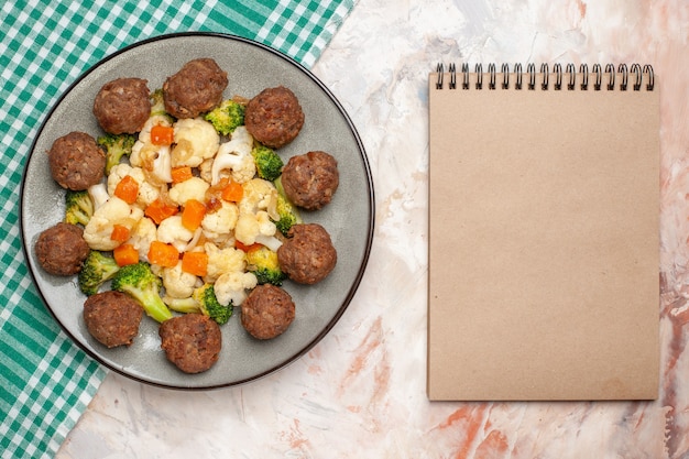 Vista superior de ensalada de brócoli y coliflor y albóndigas en un plato servilleta a cuadros verde y blanco un cuaderno sobre fondo aislado desnudo