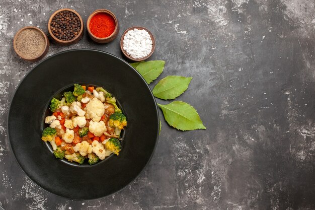 Vista superior de ensalada de brócoli y coliflor en tazón negro diferentes especias en tazones hojas sobre una superficie oscura con copia sapce